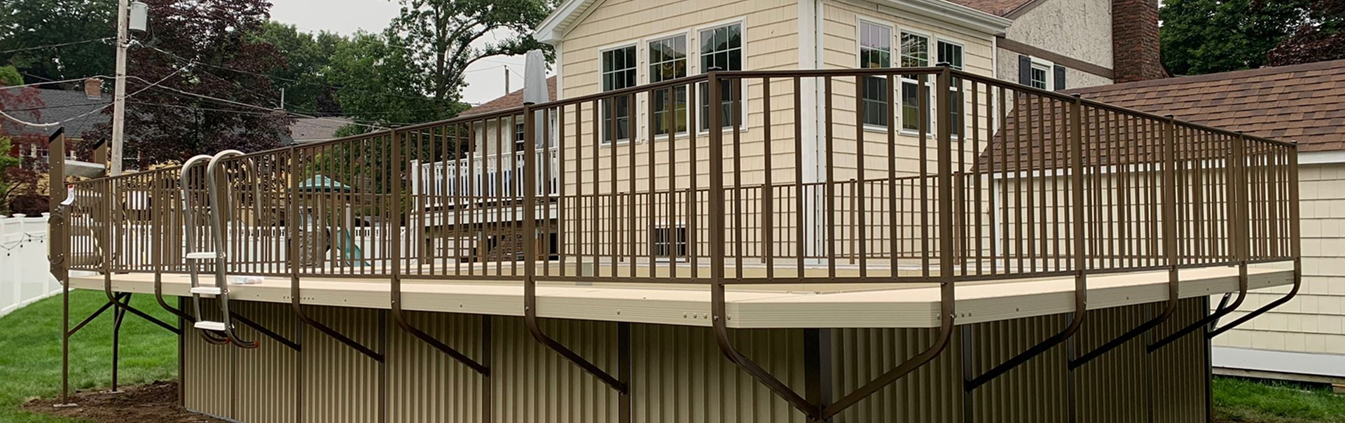 A bright suburban backyard with a swimming pool, patio seating, and a white house under clear skies.