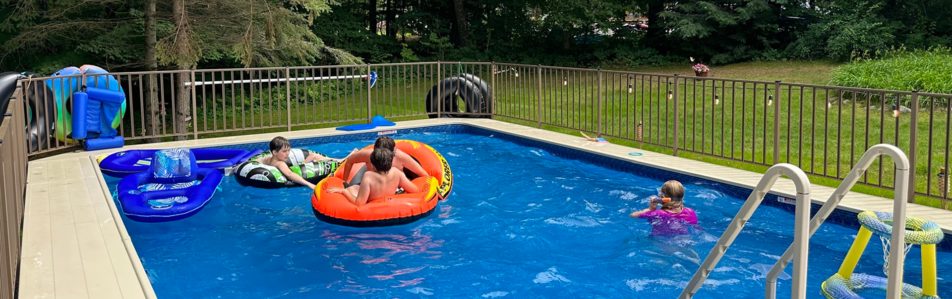 A bright suburban backyard with a swimming pool, patio seating, and a white house under clear skies.